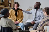 Therapist pats a man on the shoulder while leading a group therapy session.