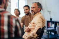 old man sits surrounded by smiling peers during a group therapy session.